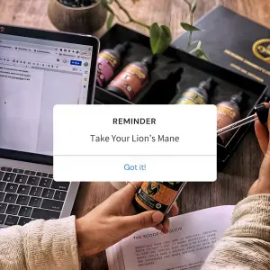 Top-down desk scene with a laptop and supplement dropper bottles, overlaid with a white 'REMINDER: Take Your Lion's Mane' calendar pop-up