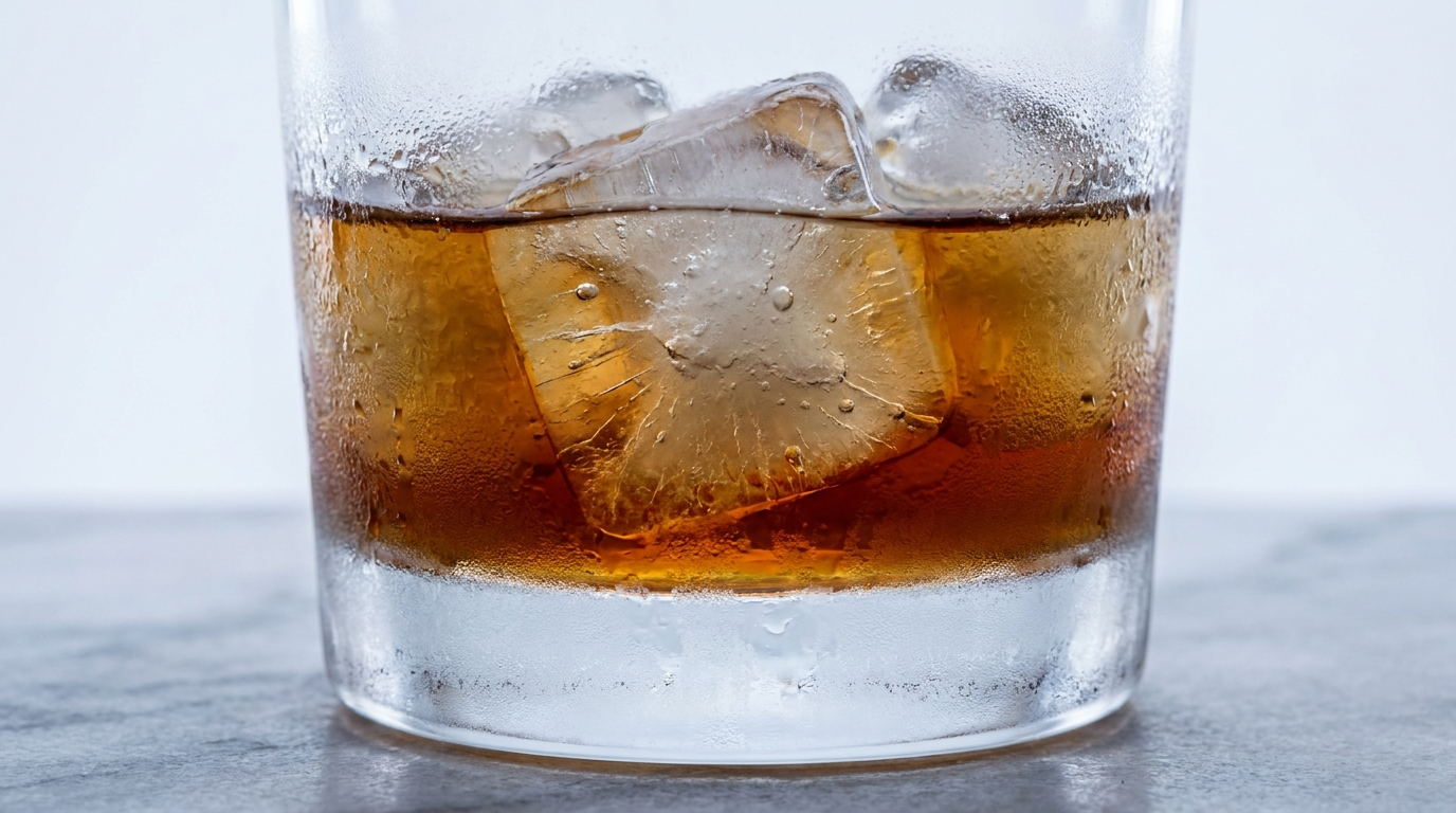 Close-up of a tumbler of amber spirit with cracked ice cubes and condensation frosting the glass