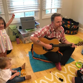 Man in a plaid shirt sitting cross-legged on a playroom floor playing acoustic guitar while young children watch