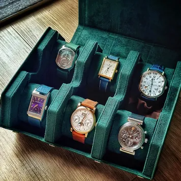 Open dark green velvet watch collector's box holding six wristwatches on a wooden table