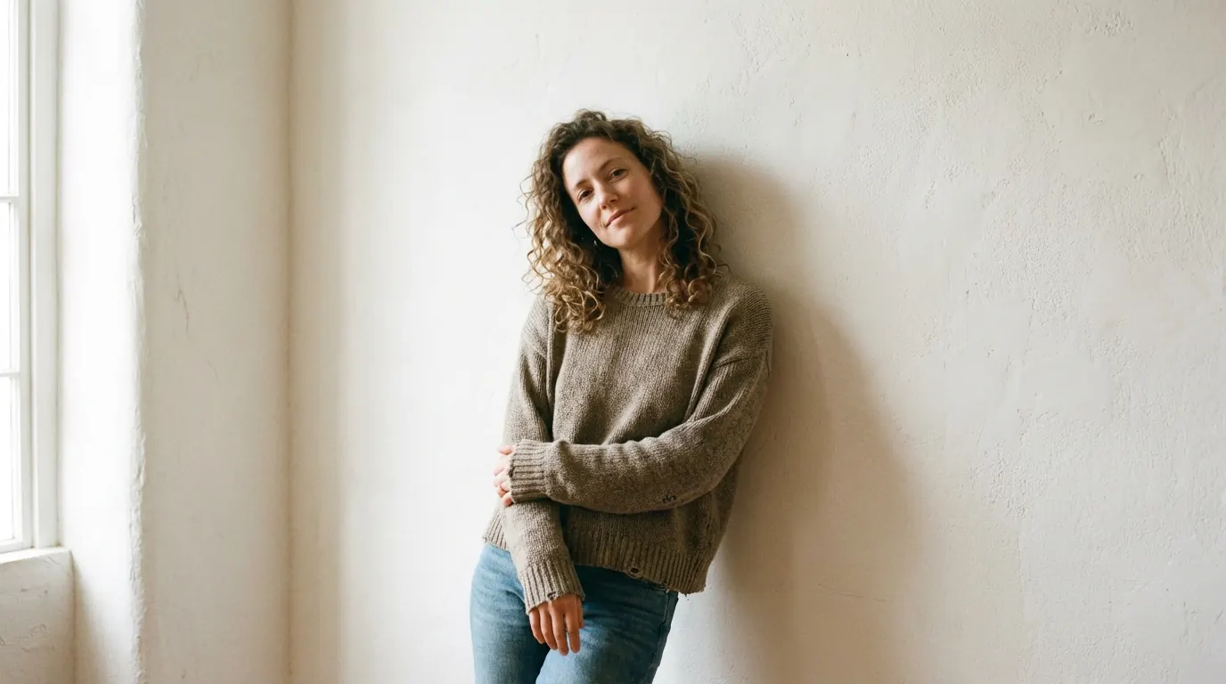 Curly-haired woman in a knit sweater leaning against a white plaster wall with arms loosely crossed